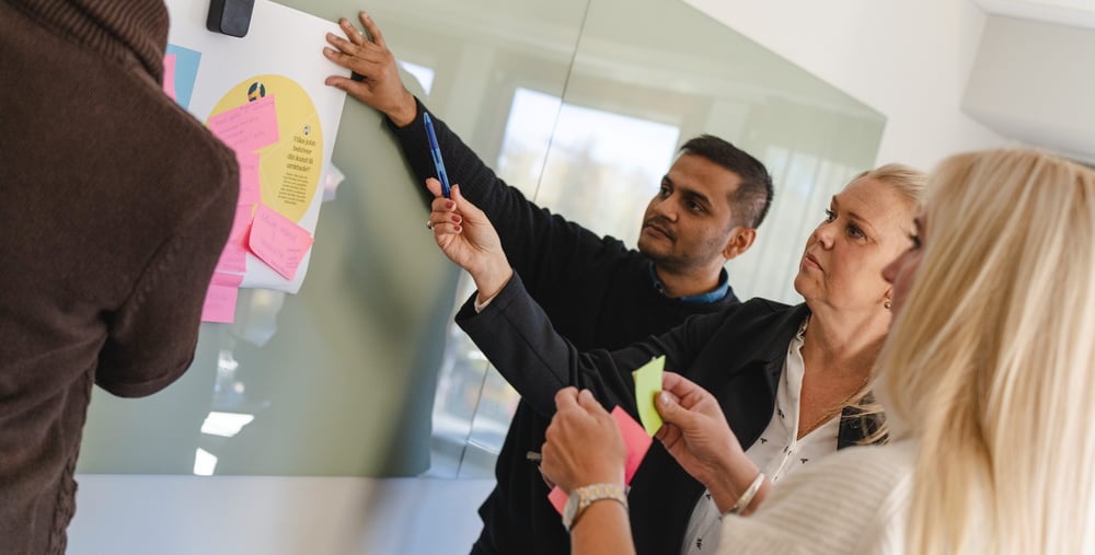 Tre personer framför en whiteboard diskuterar och sätter upp post-it lappar