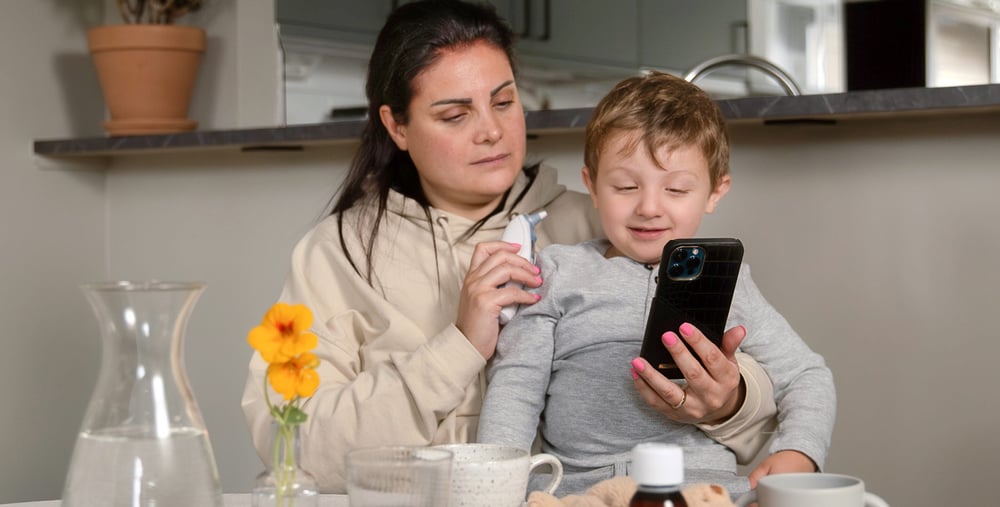 Mamma med barn i knä håller i telefon och termometer