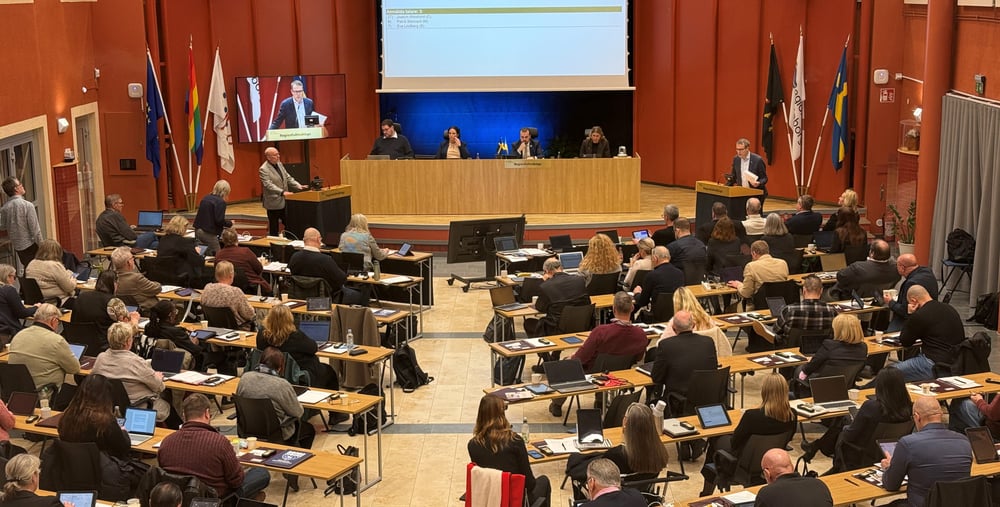En fullsatt regionfullmäktigesal med deltagare vid bärbara datorer och dokument på borden. En person talar vid en talarstol, och en presentation visas på en stor skärm med text och en talartidräknare. Jan Lahenkorva och Patrik Stenvard debatterar.