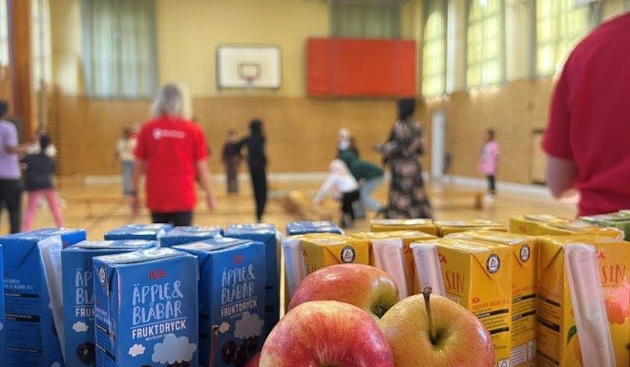 Äpplen och festisförpackningar i förgrunden, lekande barn i bakgrunden i en gymnastiksal