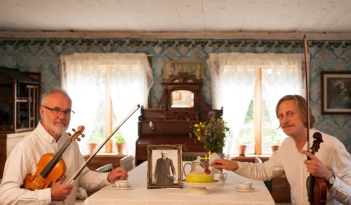 Två personer sitter vid ett bord med varsitt instrument och tittar in i kameran.