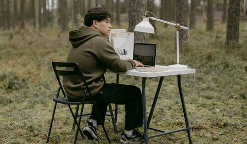 En person sitter vid ett skrivbord i skogen med dator och skrivbordslampa.