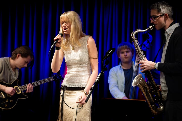 Musiker uppträder på scen med sång, gitarr, kontrabas och saxofon.